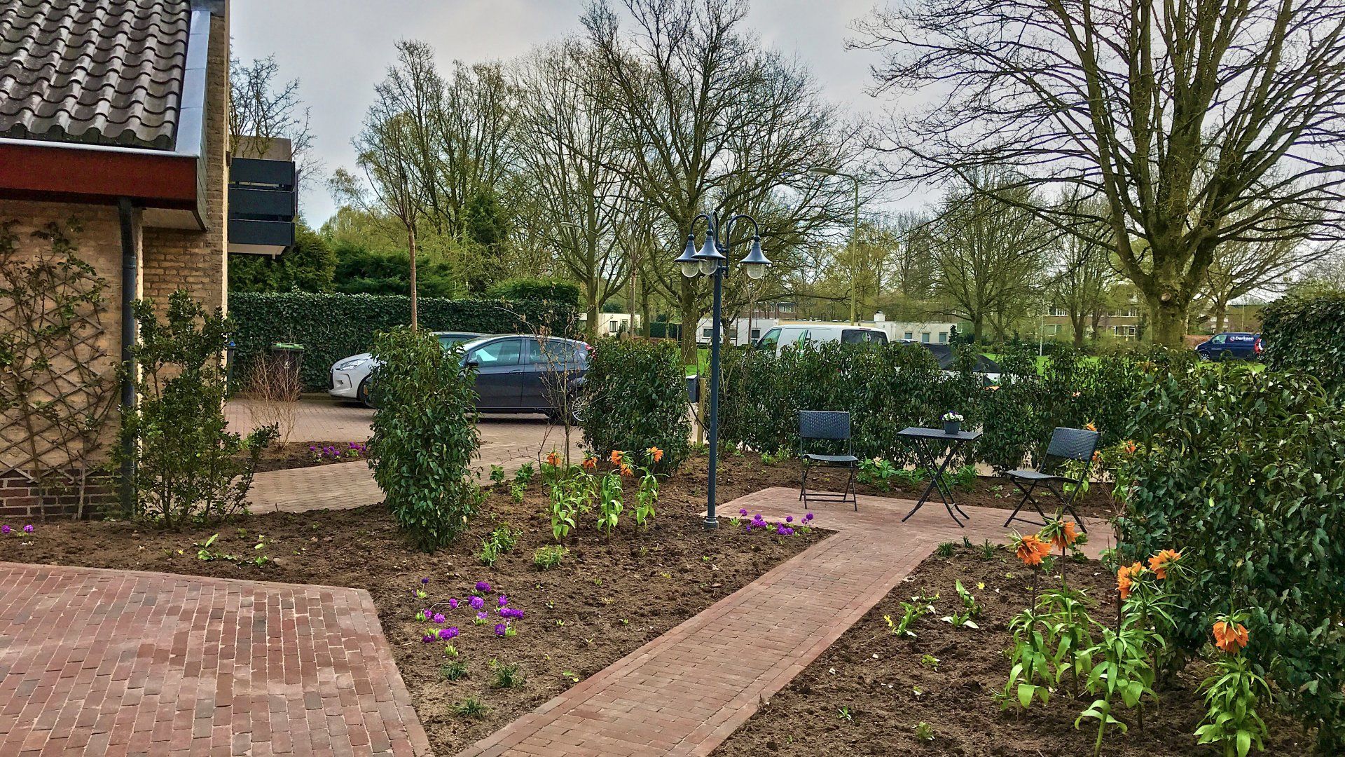 Een bakstenen pad leidt door een tuin met bloeiende planten en hagen naar een parkeerplaats.
