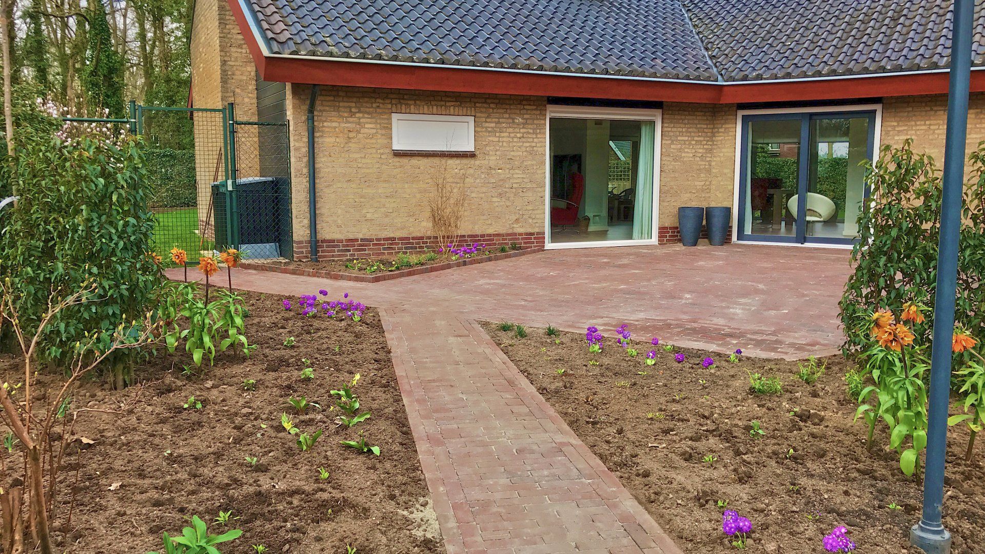 Een rood bakstenen pad leidt naar een gebouw met glazen schuifdeuren, omgeven door bloemperken.