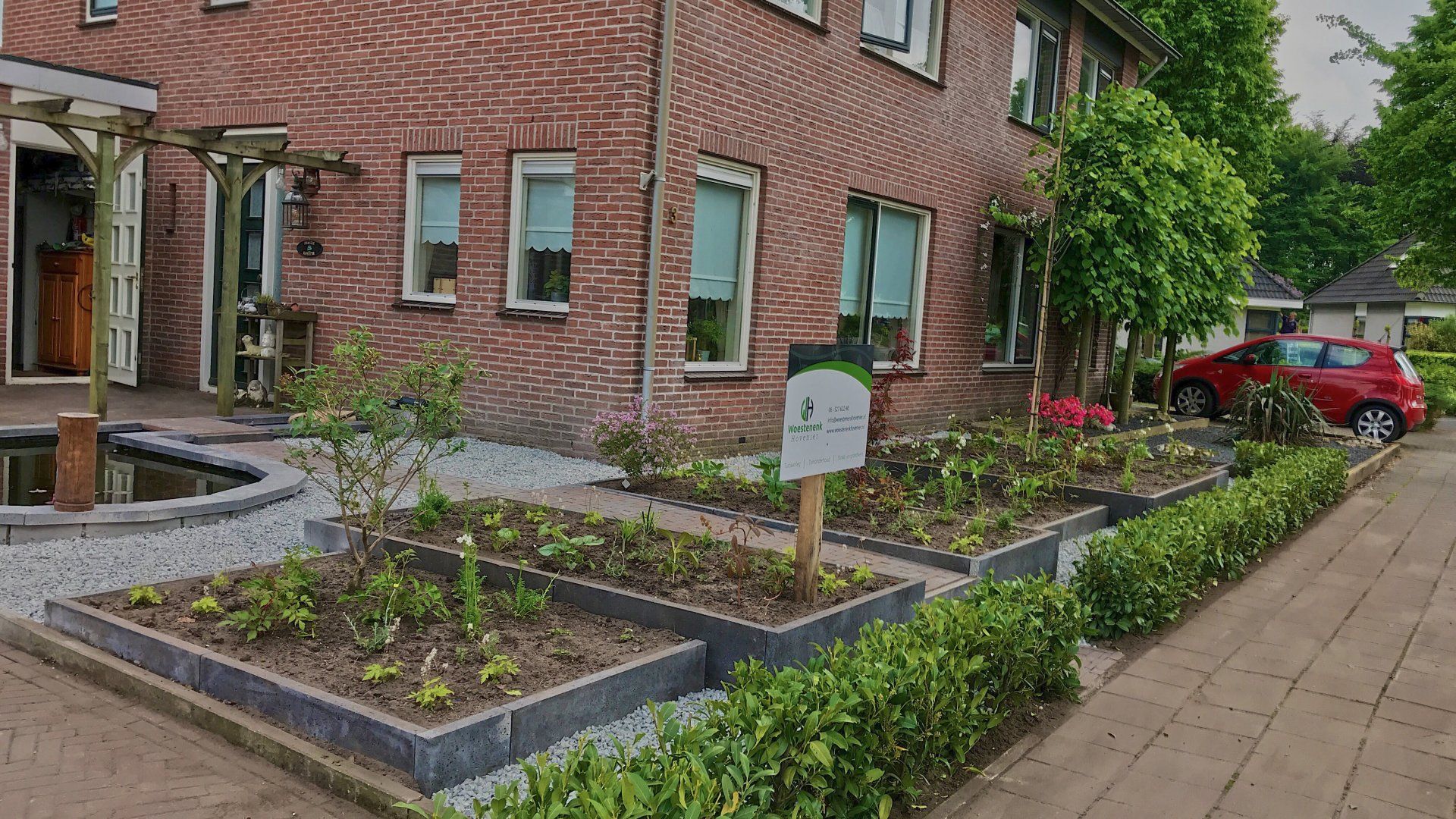 Verhoogde bloembedden met groen voor een bakstenen gebouw. ​​Een rode auto staat er vlakbij geparkeerd.