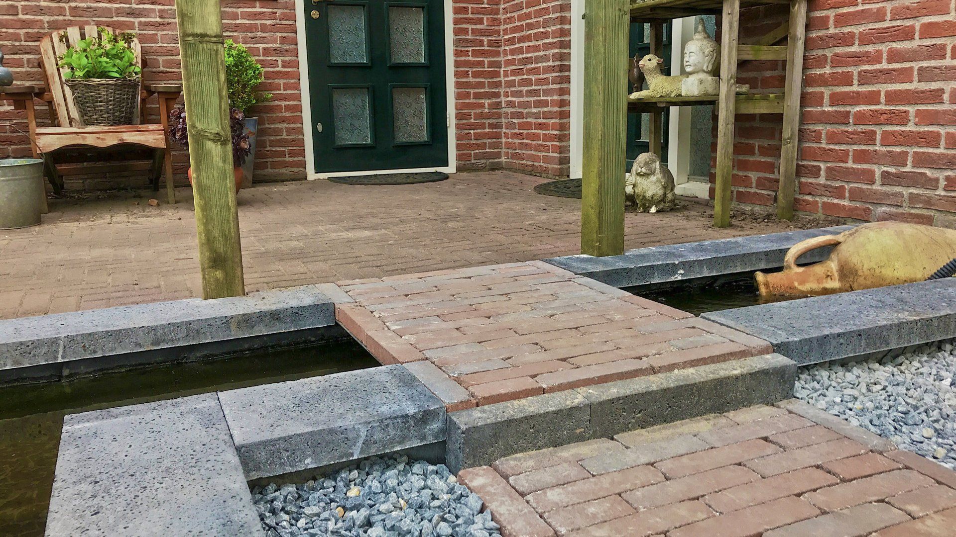 Een stenen terras met een waterpartij, stenen randen en een brug die naar een groene deur leidt.