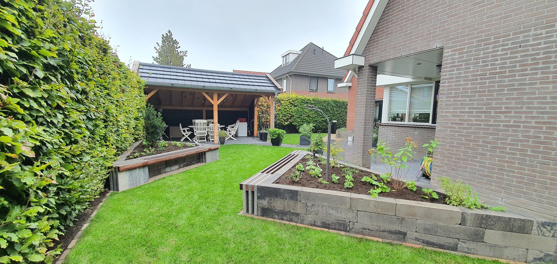 Een achtertuin met een weelderig groen gazon, bloemperken en een overdekt terras.