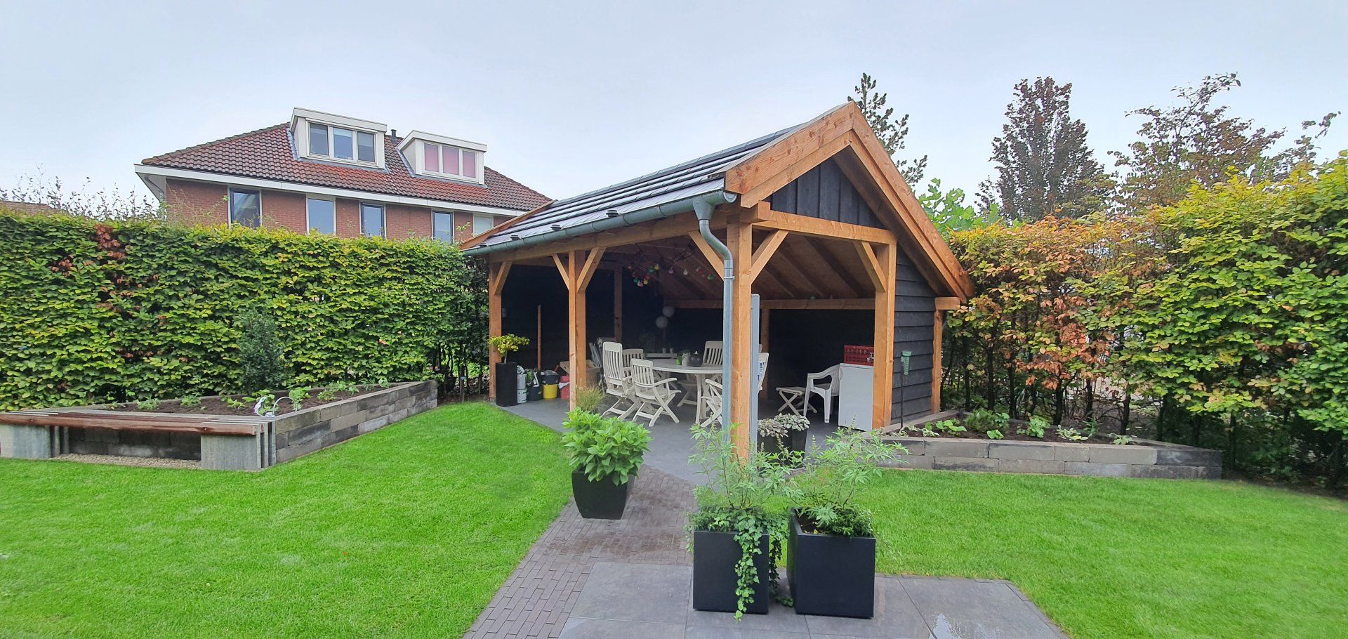 Een houten tuinhuisje met een tafel en stoelen, omgeven door groen en een gazon.