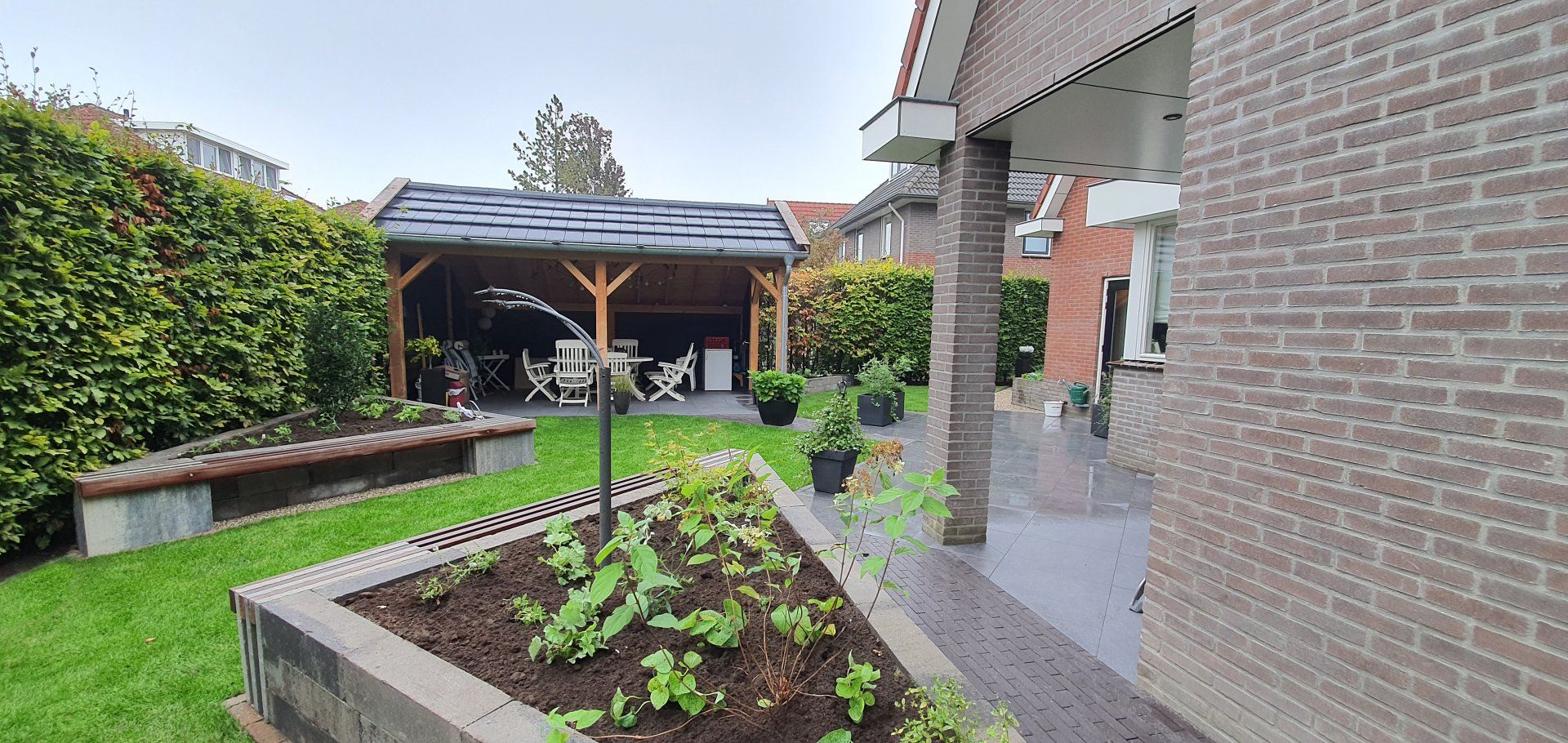 Achtertuin met een bakstenen huis, houten terras, tuinhuisje, groen gazon en bloembedden. Bewolkte lucht.