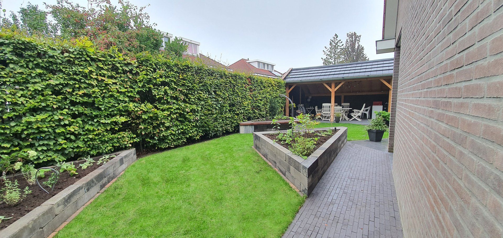 Een weelderige achtertuin met een groen gazon, verhoogde bloembedden, een overdekt terras en een bakstenen huis.