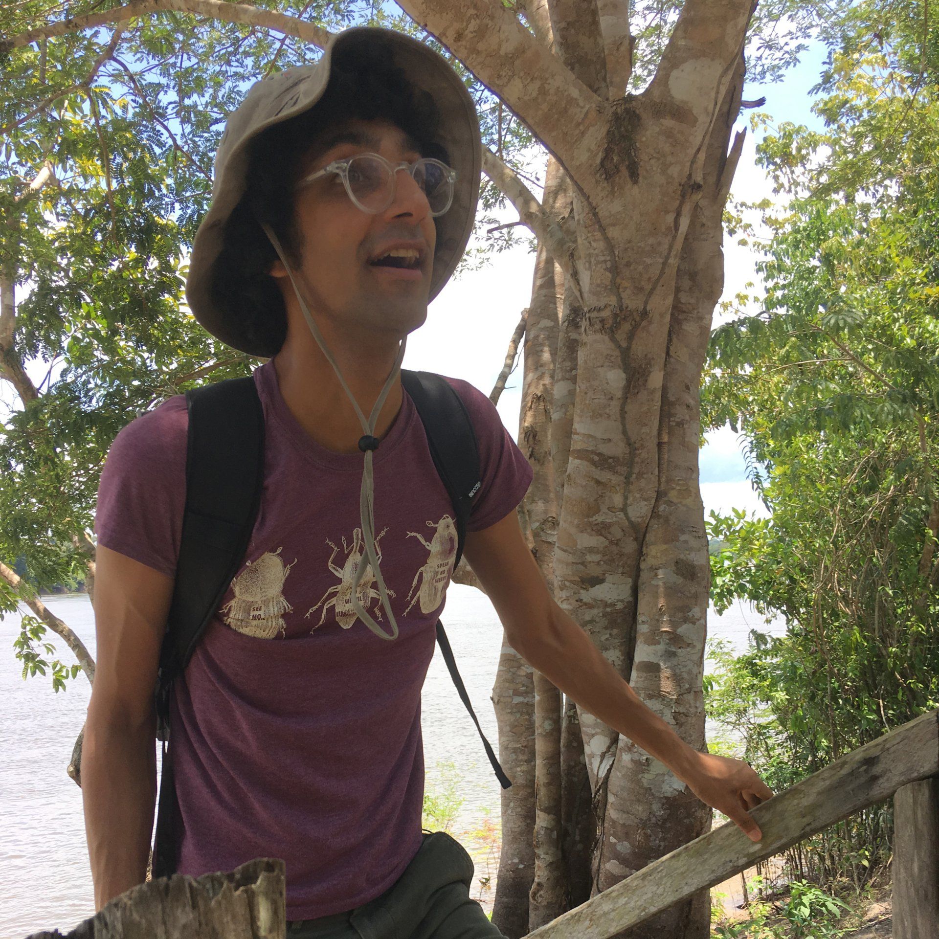 A man wearing a purple shirt and a hat is standing next to a tree