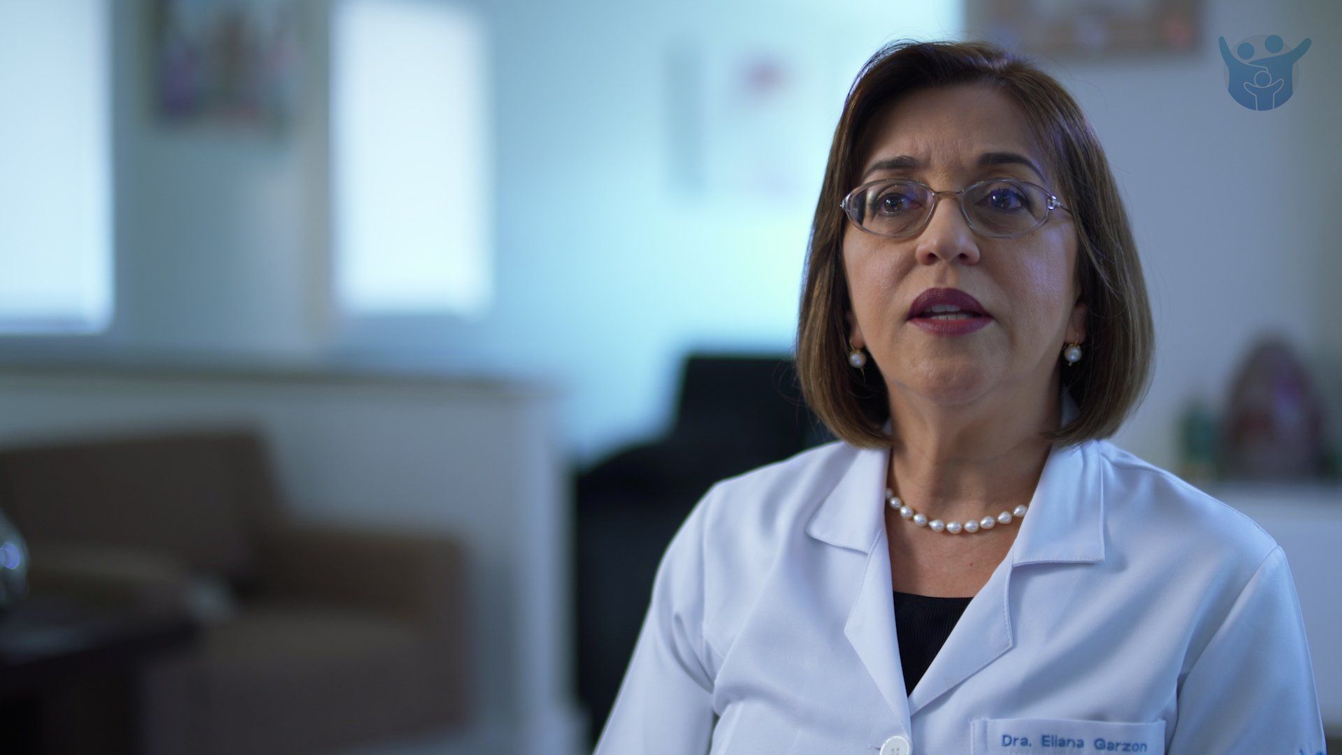 A woman in a lab coat and glasses is talking to a camera.