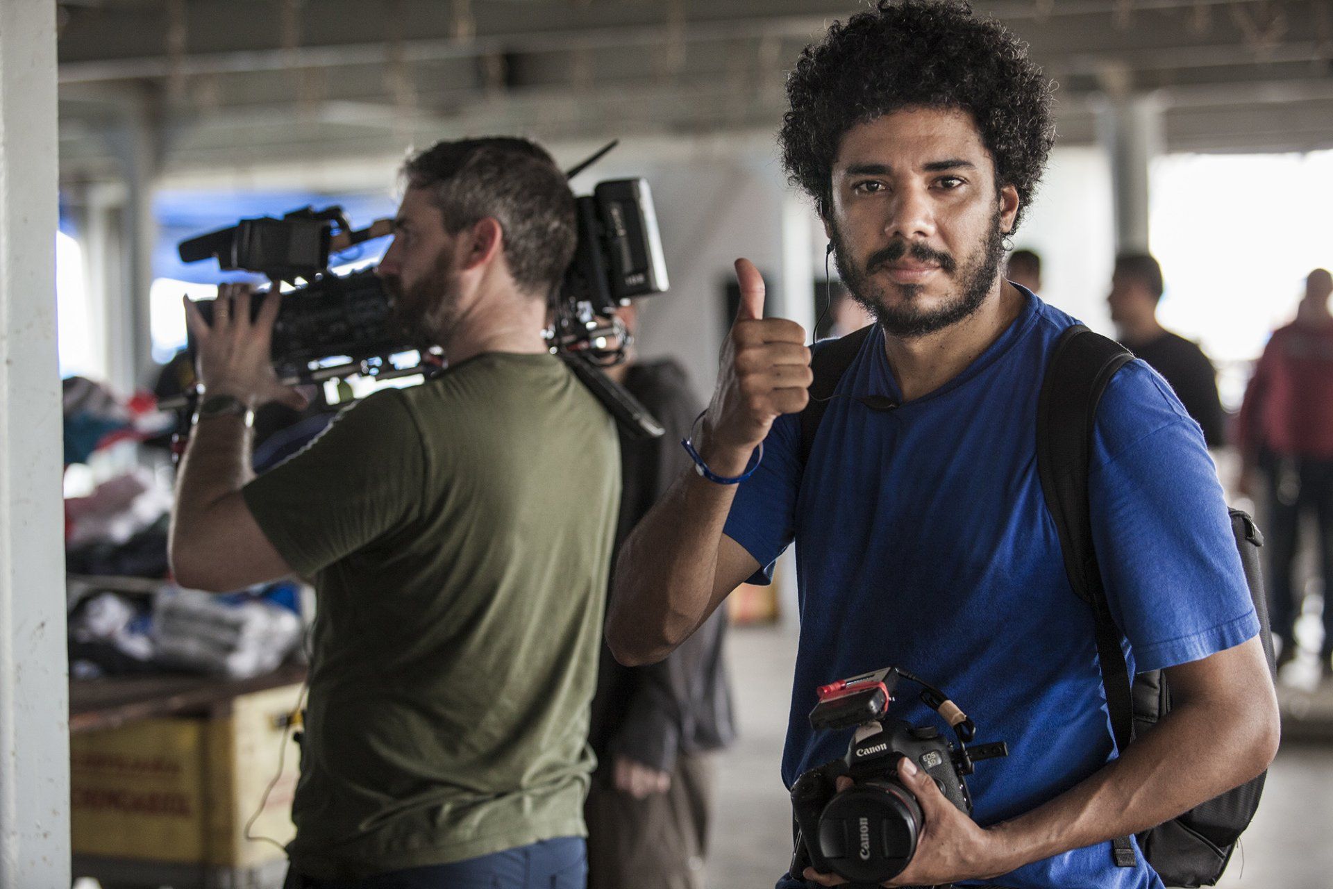 A man in a blue shirt is holding a camera and giving a thumbs up.