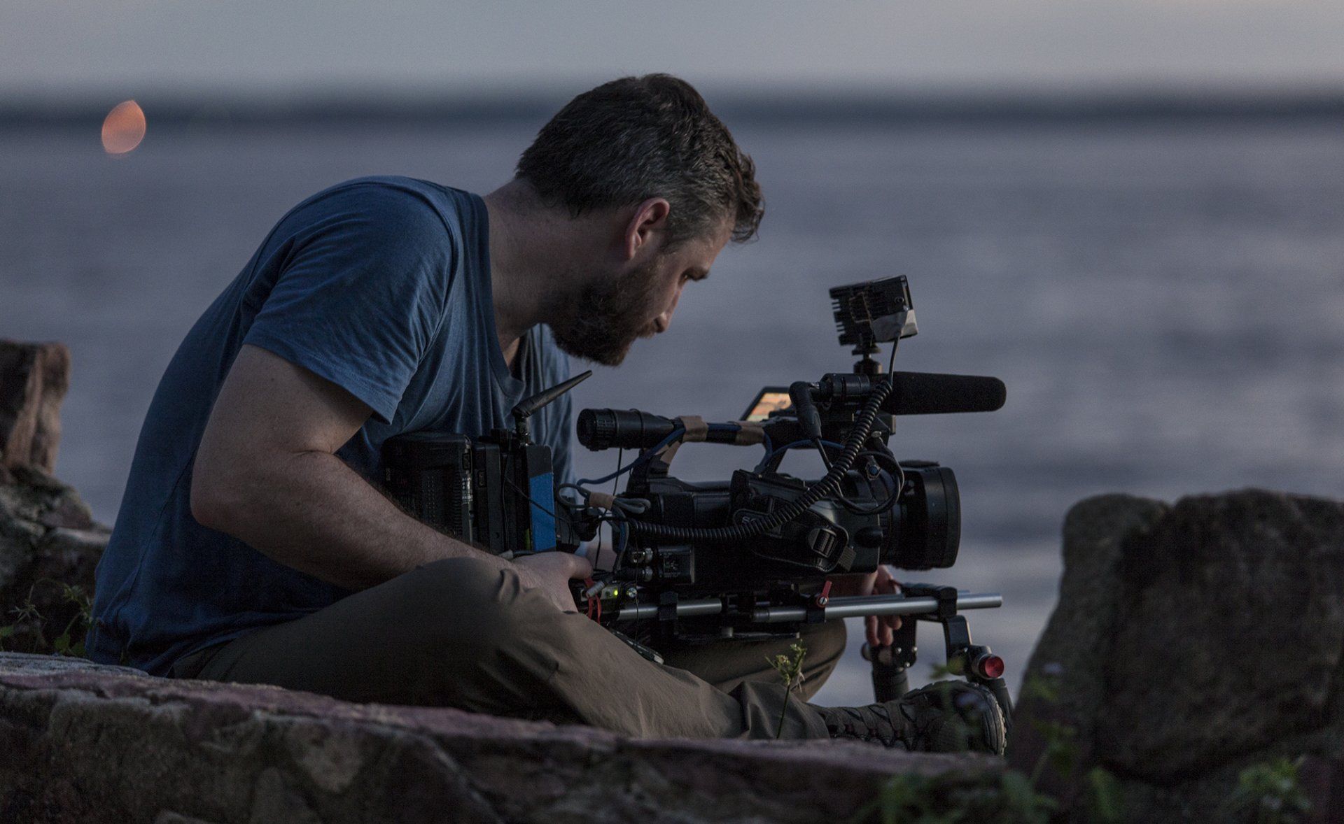 A man is sitting on a rock holding a camera in front of the ocean.