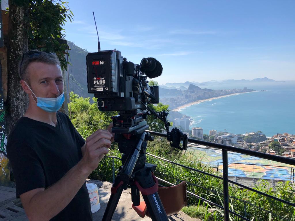 A man wearing a mask is standing in front of a camera on a tripod.