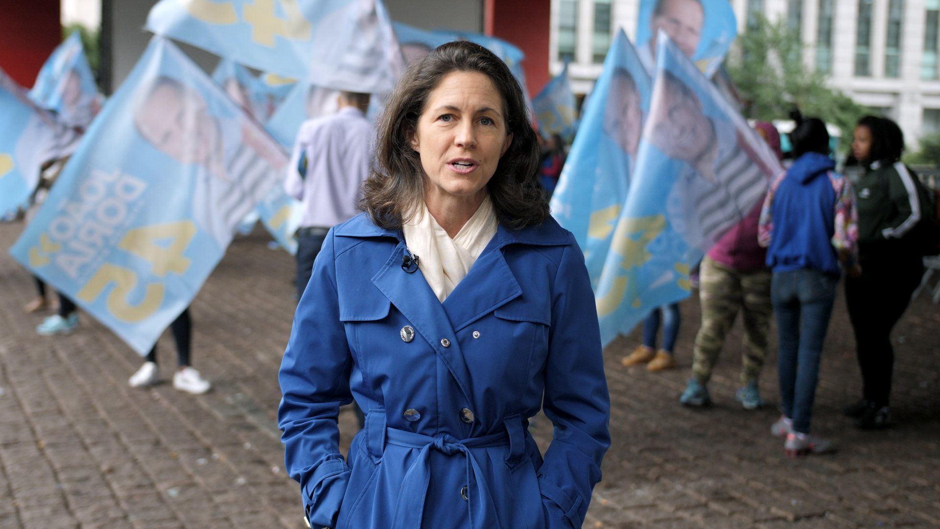 A woman in a blue coat is standing in front of a crowd of people holding blue flags.