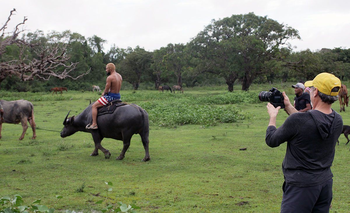 A man is riding a water buffalo in a field while a woman takes a picture.