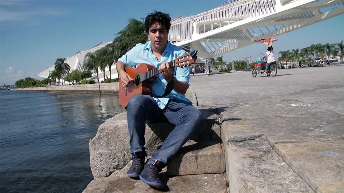 A man is sitting on a rock playing a guitar by the water.