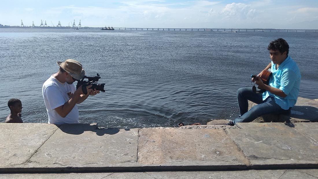Two men are sitting on a concrete wall near the water taking pictures.
