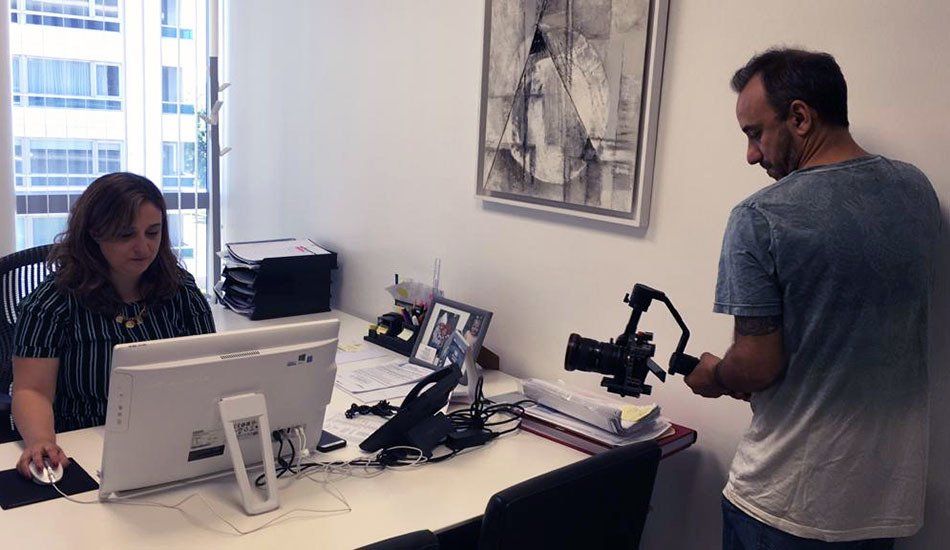 A man is taking a picture of a woman sitting at a desk with a computer.