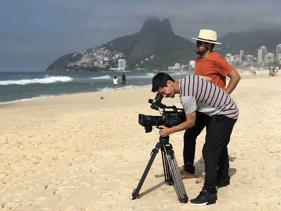 Two men are standing on a beach looking at a camera.