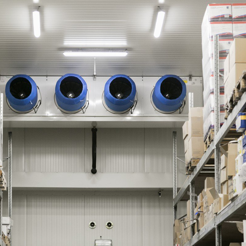 Grote blauwe ventilatoren zijn hoog in een magazijngang gemonteerd, naast de schappen met producten.