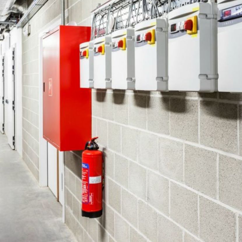 Brandveiligheidsapparatuur gemonteerd aan een betonnen muur in een gang, waaronder rode kasten en een brandblusser.