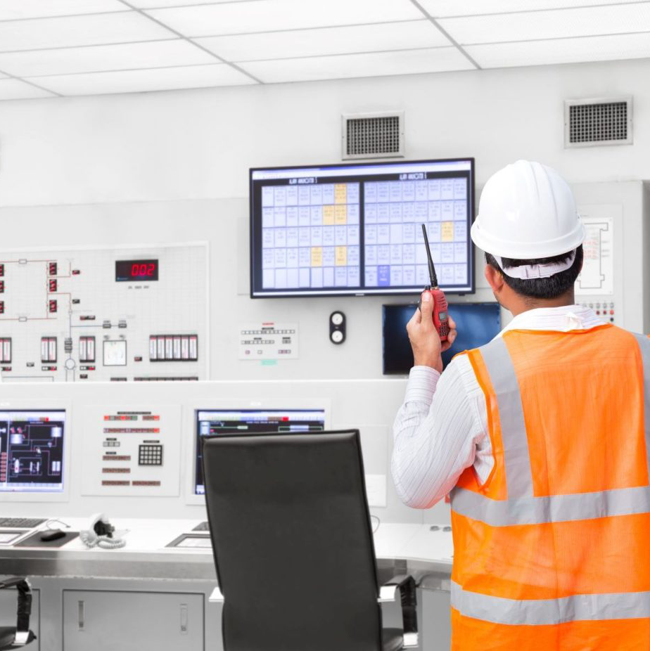Technicus in oranje veiligheidsvest en witte helm bekijkt bedieningsschermen in een industriële controlekamer.