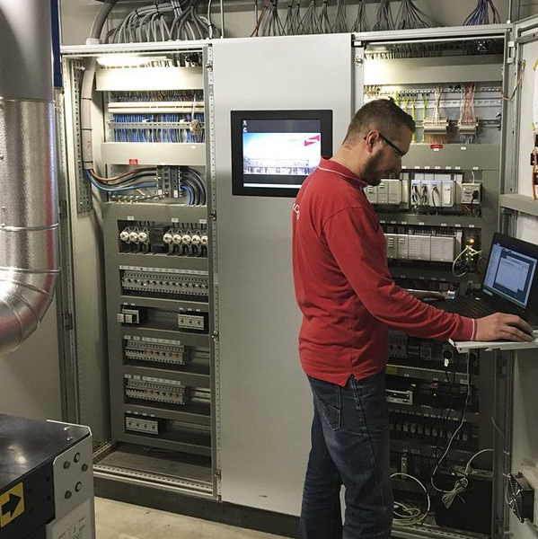 Een technicus in een rood shirt werkt aan een laptop naast een wand met schakelkasten voor elektrische apparatuur.