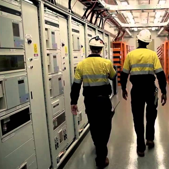 Twee werknemers met veiligheidshelmen lopen door een smalle gang van een industriële controlekamer.