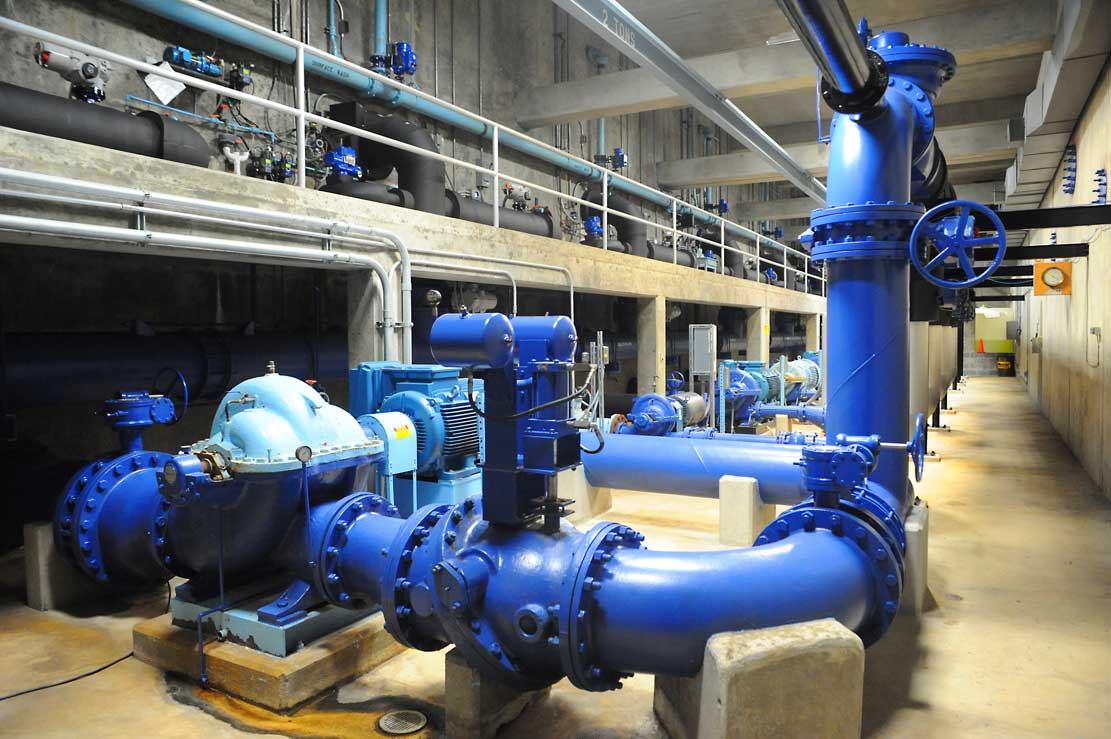 Blauwe industriële leidingen en pompen in een technische ruimte van een fabriek met loopbruggen erboven.