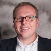 Lachende man met bril en donkere blazer tegen een grijze studioachtergrond.
