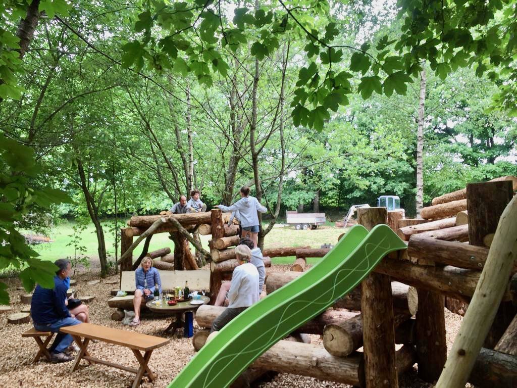 Een groep mensen zit aan een picknicktafel in het bos, vlak bij een glijbaan.