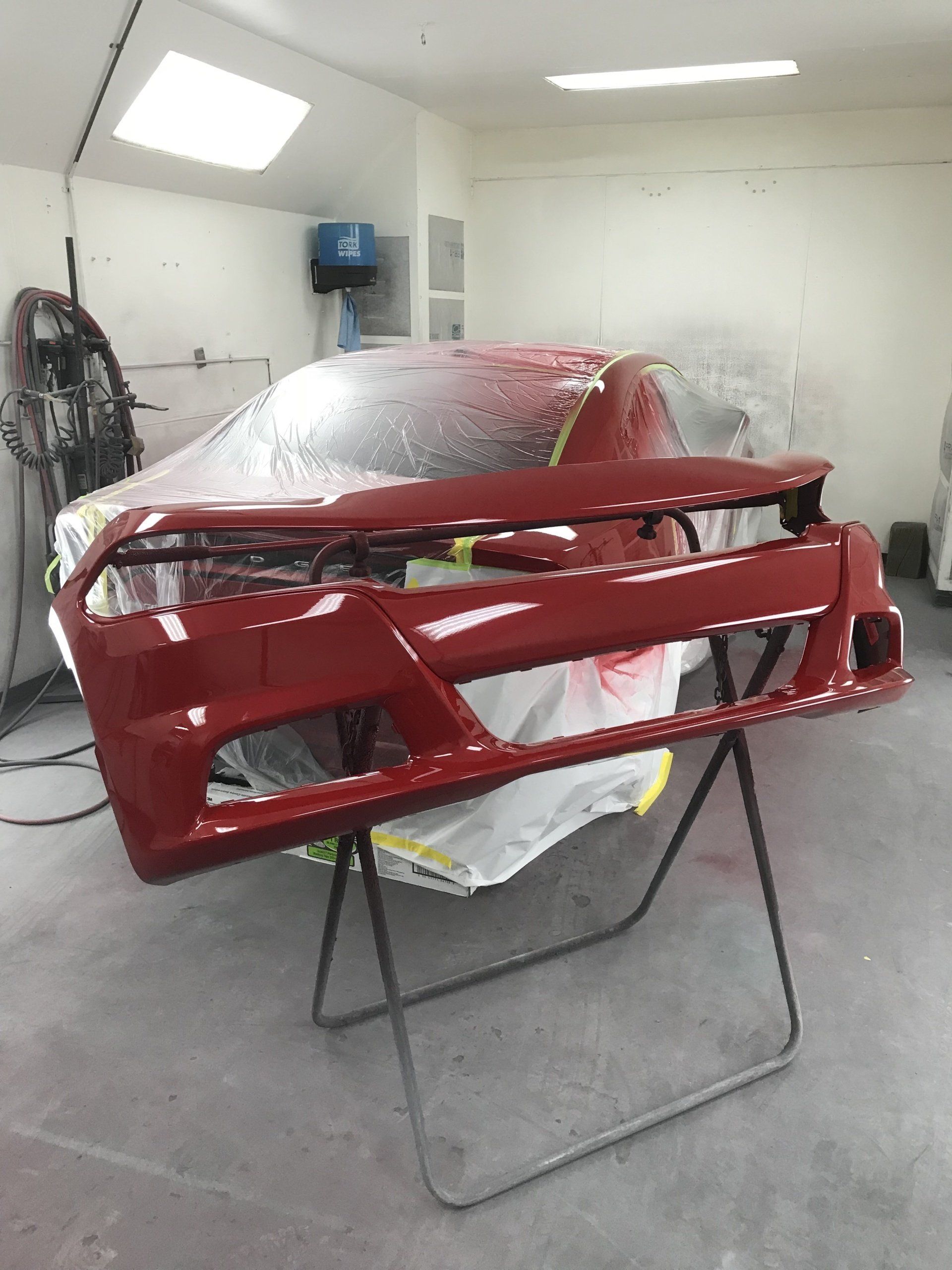 Red car bumper on a stand in a paint shop, car masked in background.