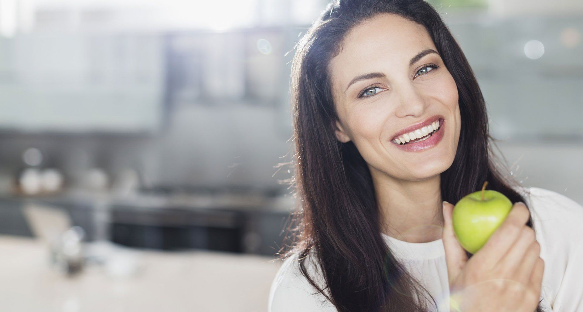 Woman smiling, holding a green apple. Long dark hair, white shirt, blurry kitchen background.