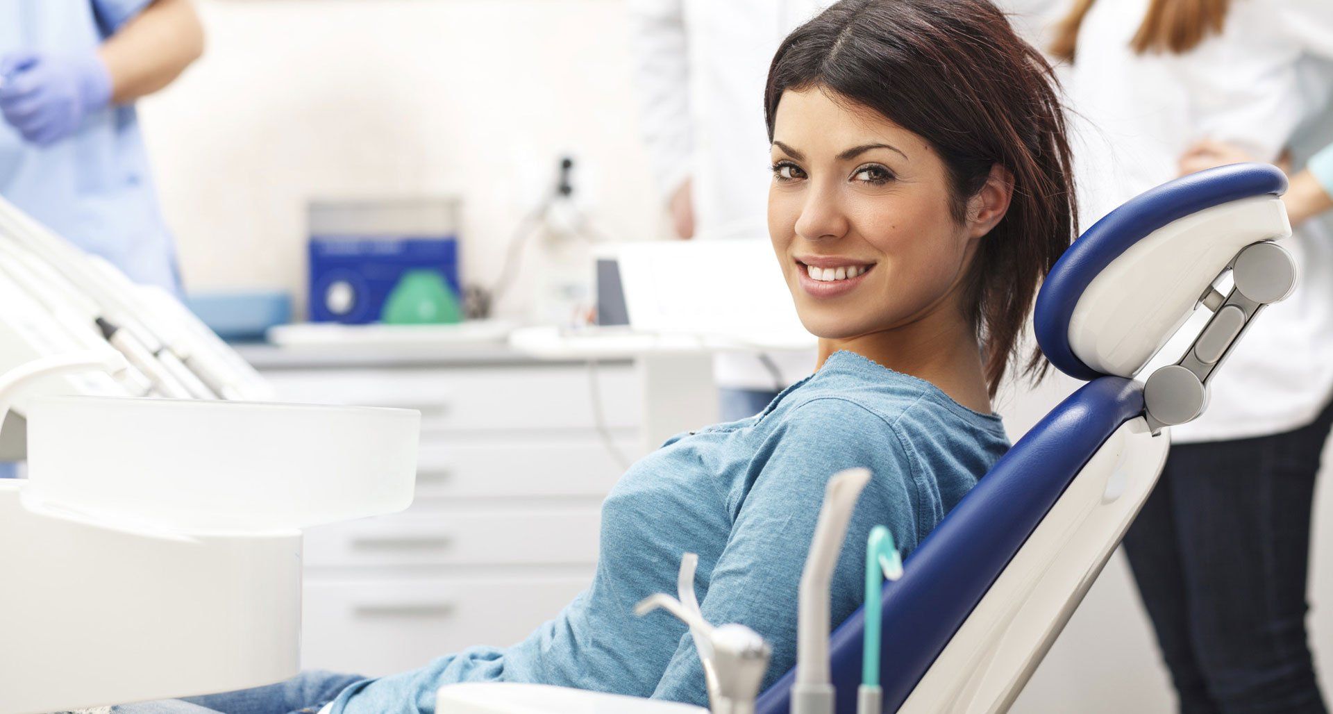 Woman in a dental chair smiles at the camera; dentist and assistant in the background.