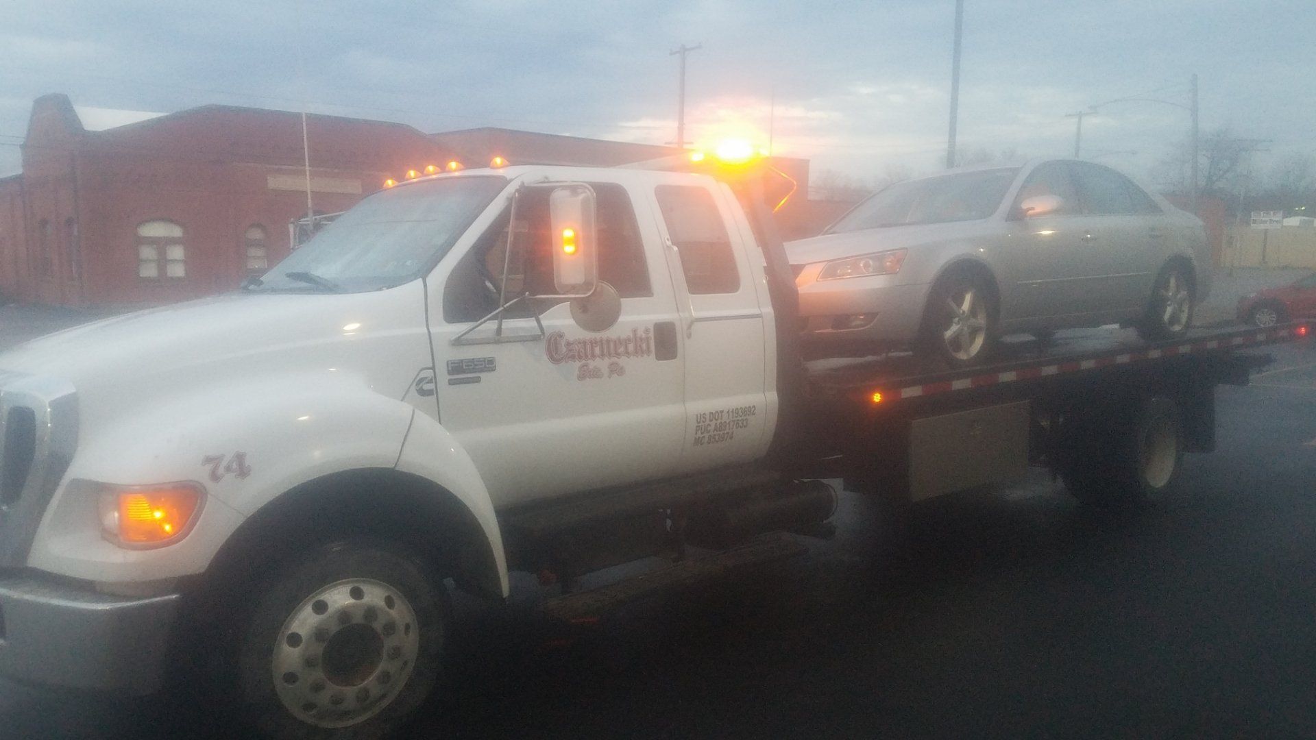Tow truck with a silver car on its flatbed, parked in front of a brick building on a cloudy day.