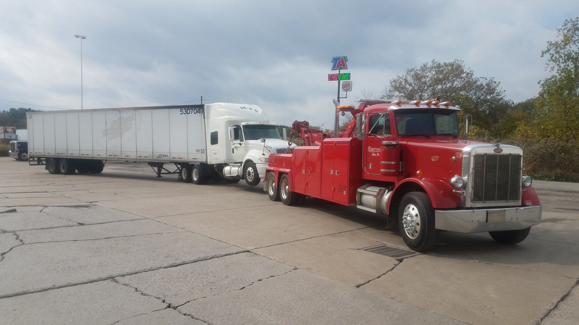 Red tow truck towing a white semi-truck and trailer on a gray paved area near a roadside sign.