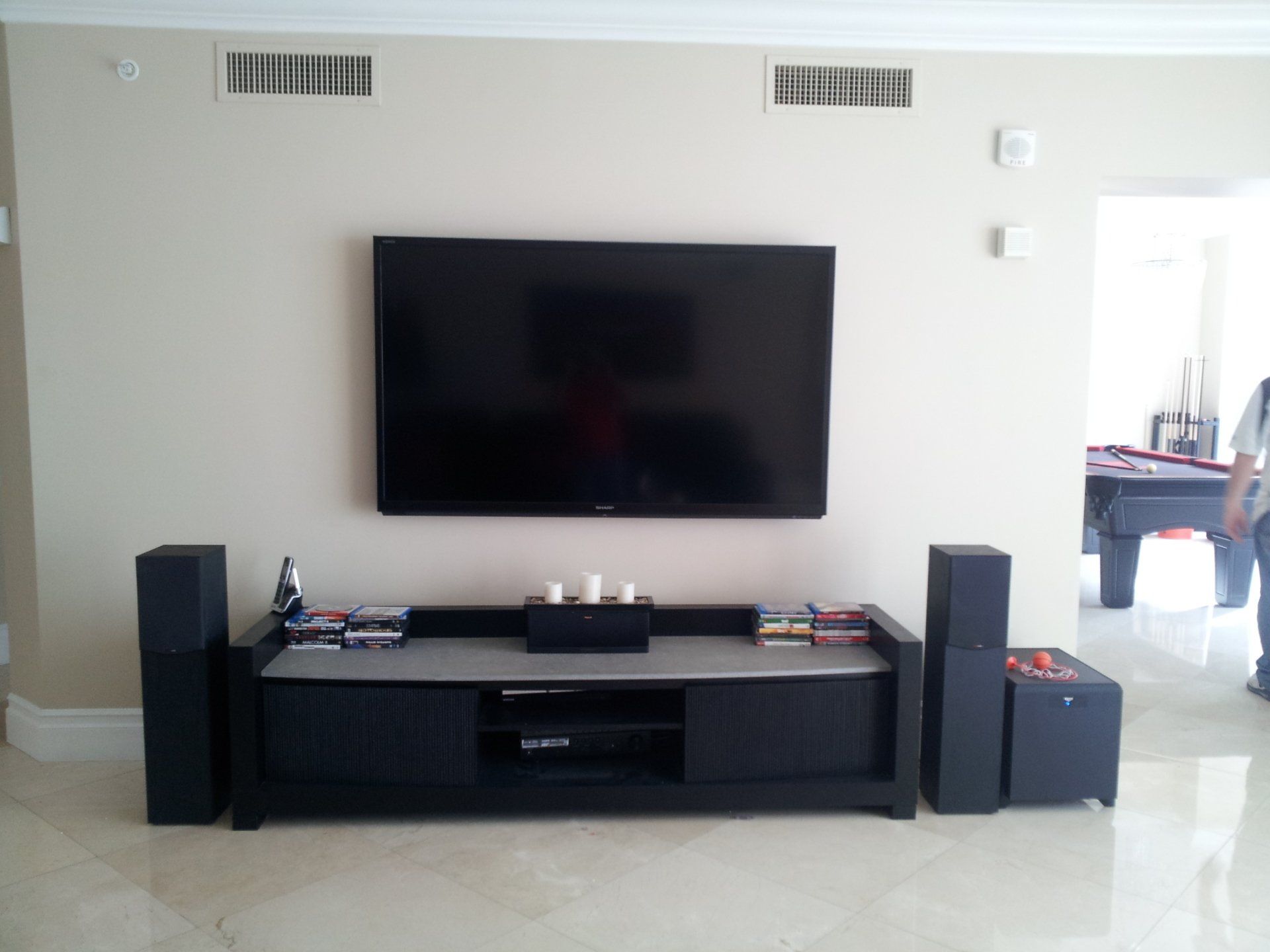 A wall-mounted TV above a black entertainment center with speakers and a subwoofer; a pool table in the background.