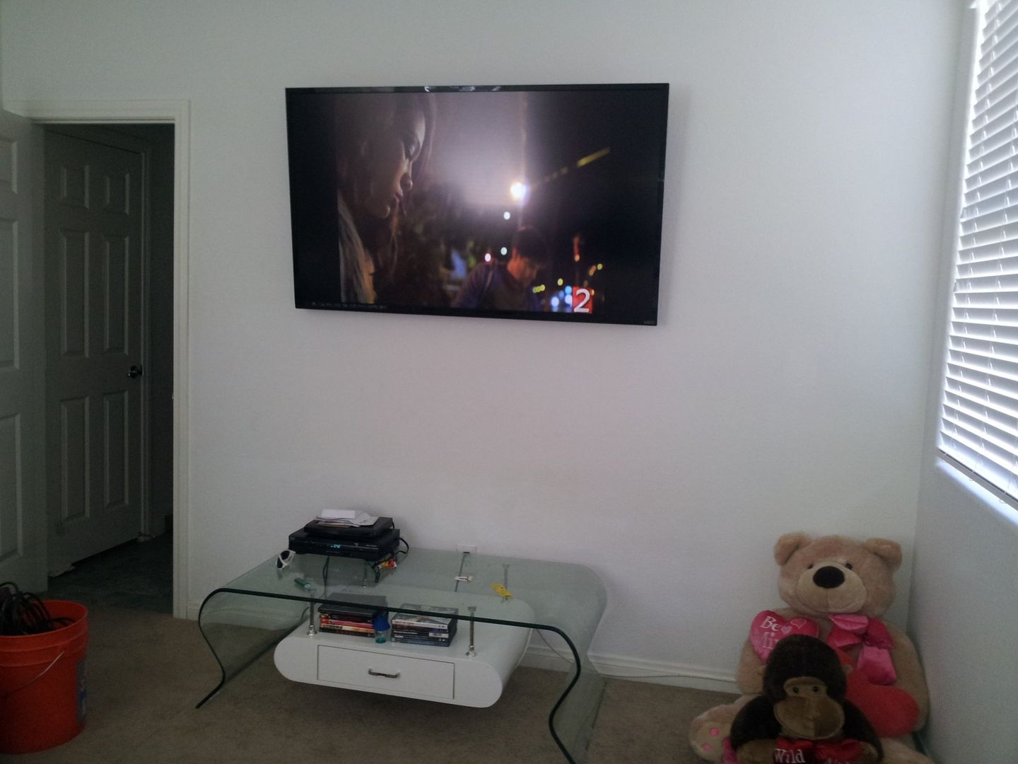 A mounted TV on a white wall, over a glass table with electronic devices and toys in a room.