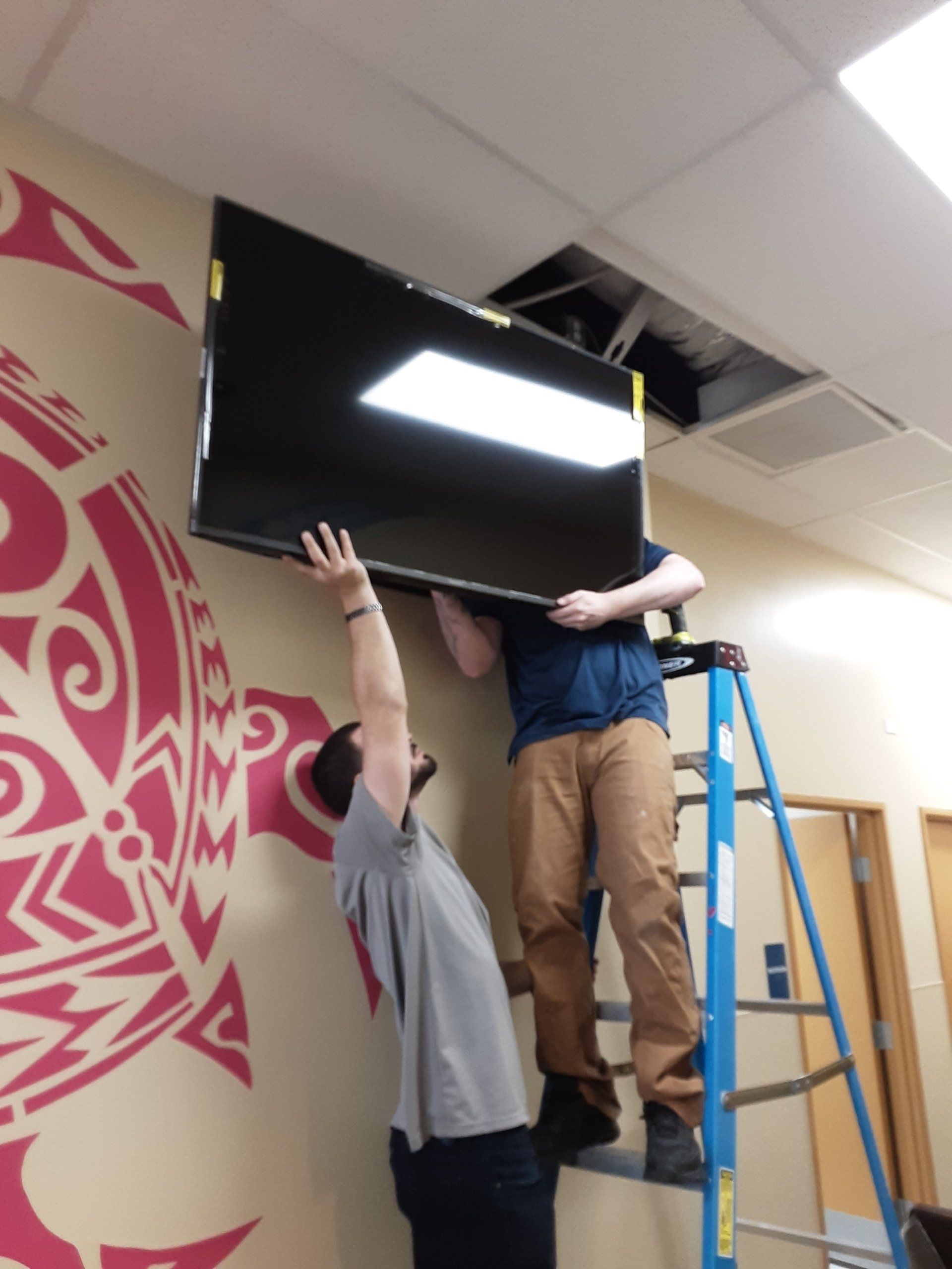 Two people install a large TV on a wall, one on a ladder, near a ceiling opening and patterned wall.