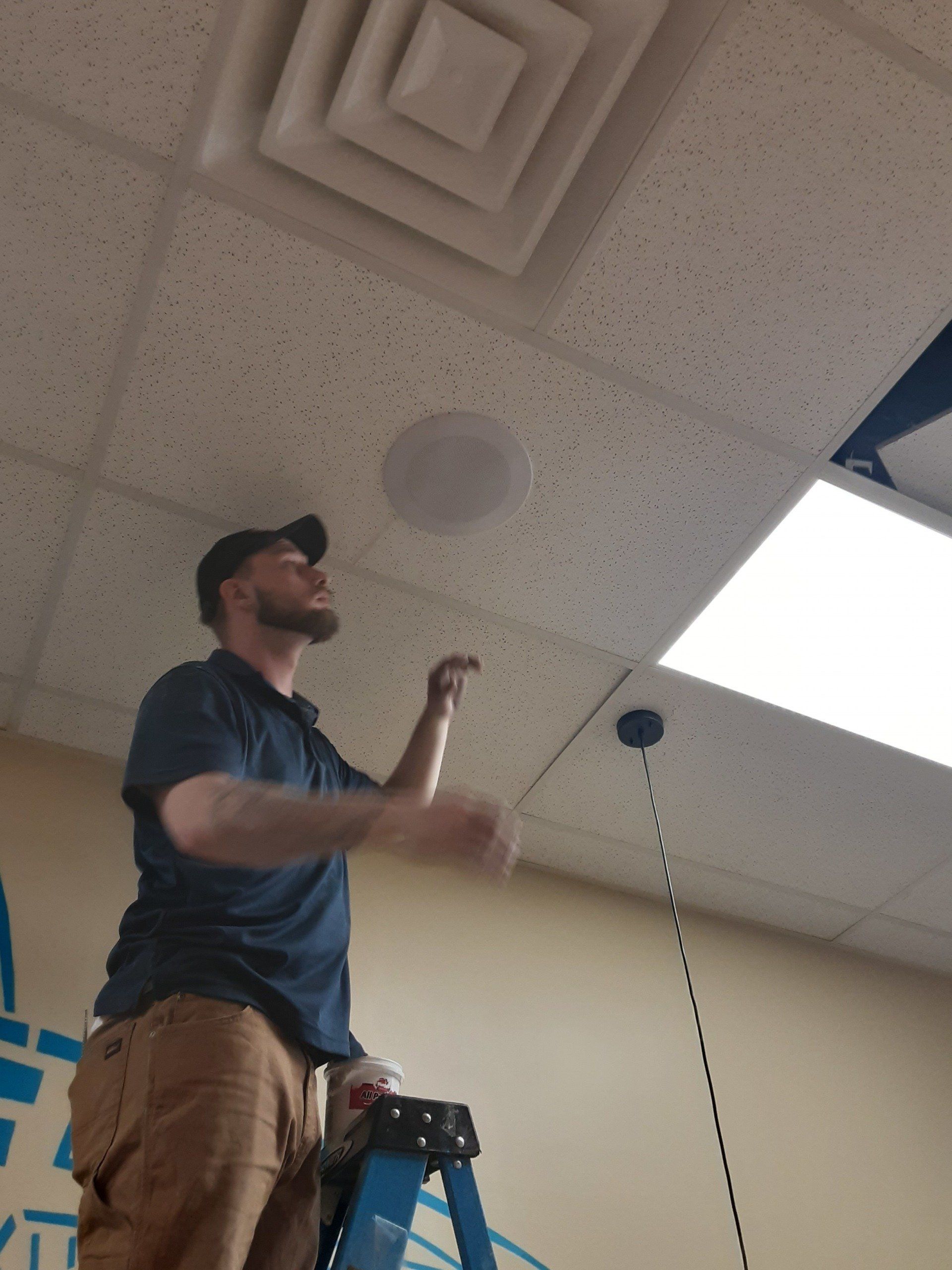 Man on a ladder installing a ceiling speaker. He's reaching up towards the speaker near an air vent.