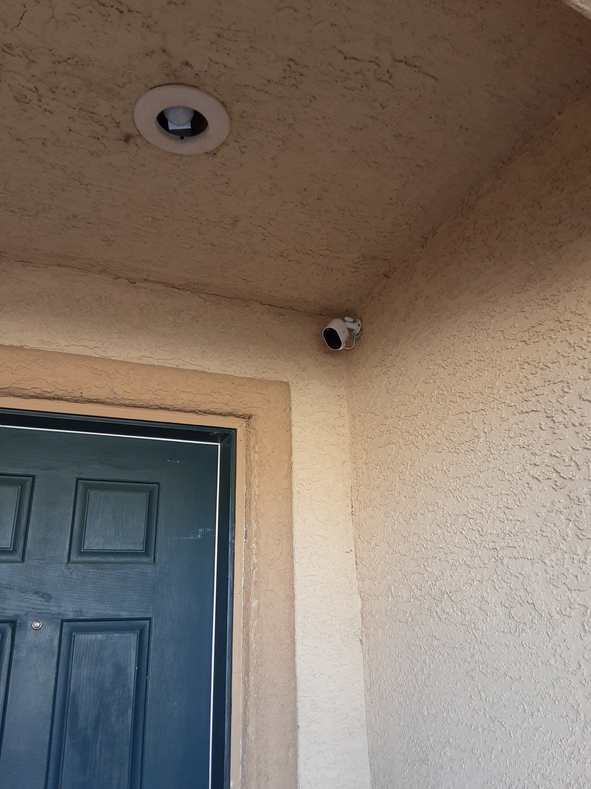 Security camera mounted on a beige stucco wall, near a recessed light and a blue door.