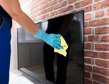 Person in blue gloves cleaning a black TV screen with a yellow cloth in front of a brick wall.
