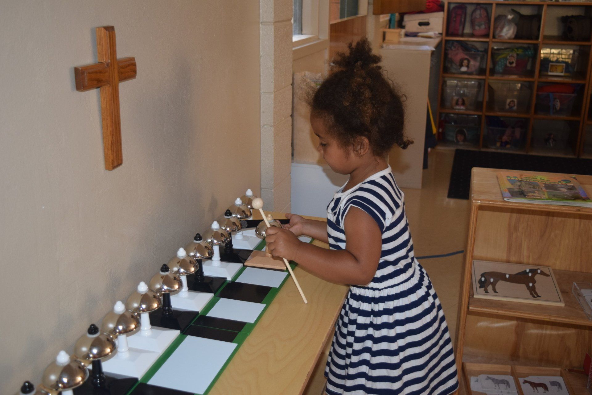 A young girl in a striped dress plays a set of bells in a room with a cross on the wall.