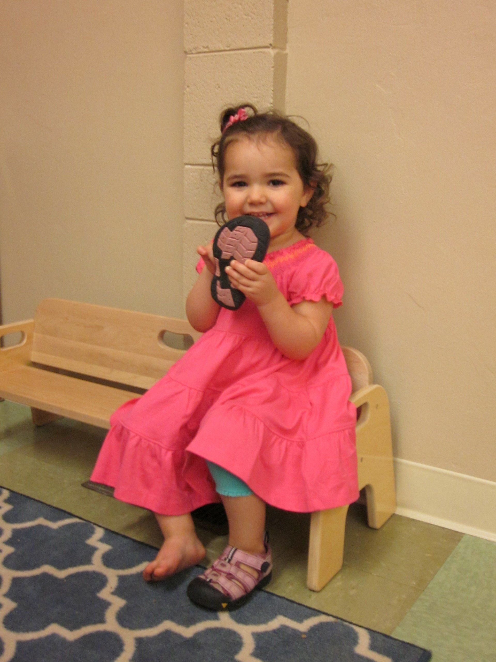 Smiling toddler in pink dress, holding sandal, sitting on bench.