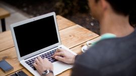 A man is typing on a laptop computer at a wooden table.