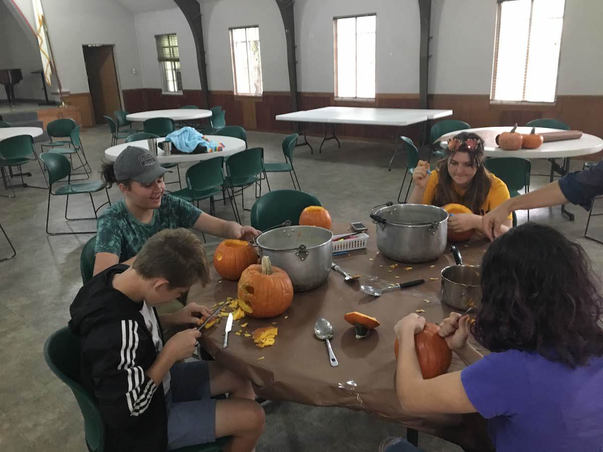 Group of teens carving pumpkins around a table in a large room with windows.