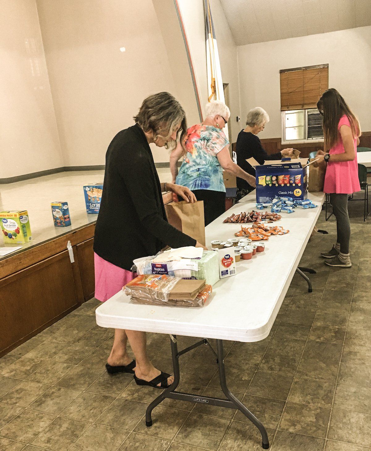 Volunteers pack food bags at a long table in a room. People are mostly older women, one teen.