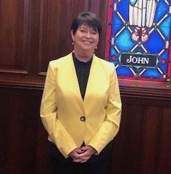 Woman with short dark hair in yellow jacket smiles near stained glass window.