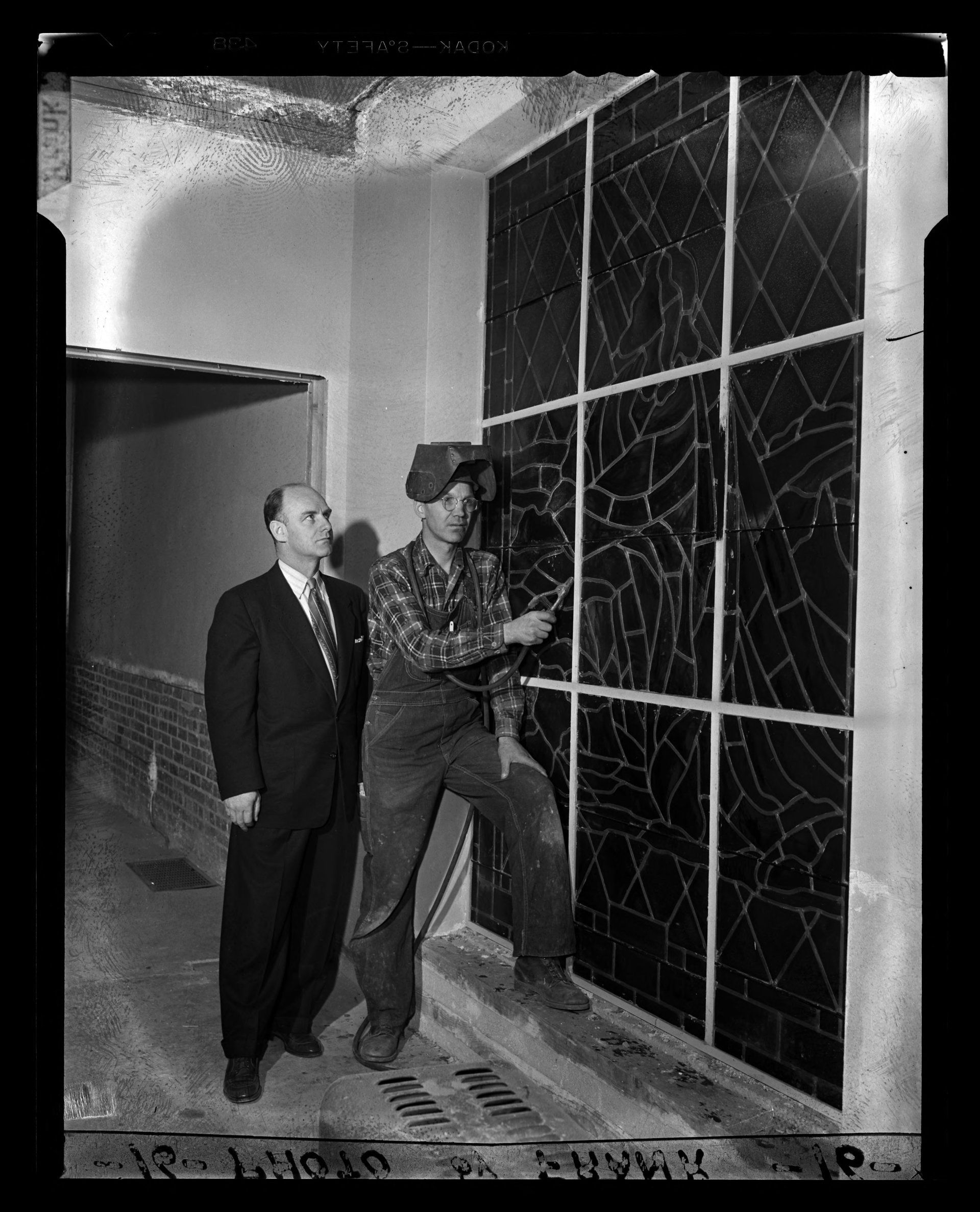 Two men examine a large, broken panel. One in a suit, the other overalls, holding a tool. Indoor setting.
