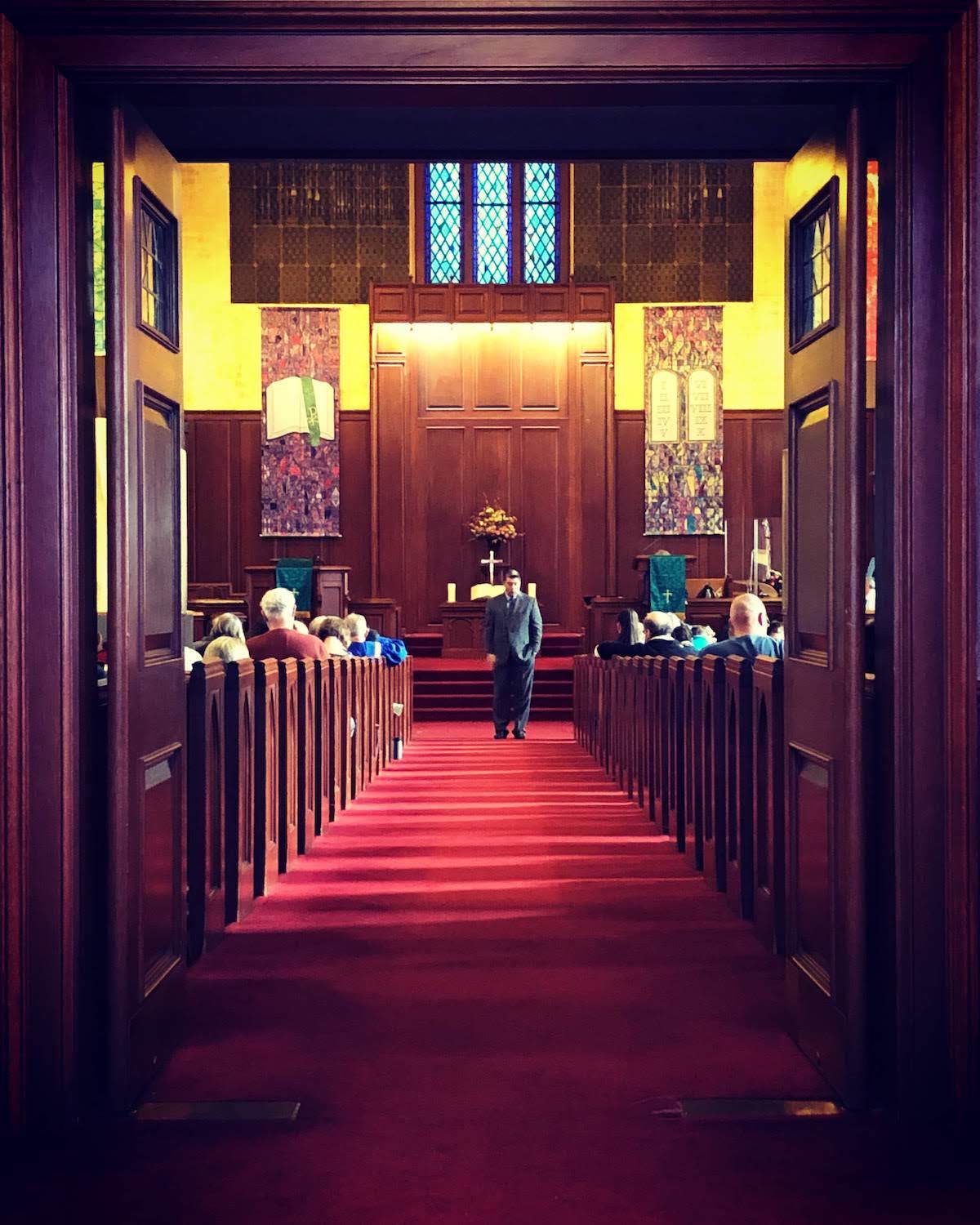 Interior of a church: rows of pews, red carpet, person walking towards the altar.