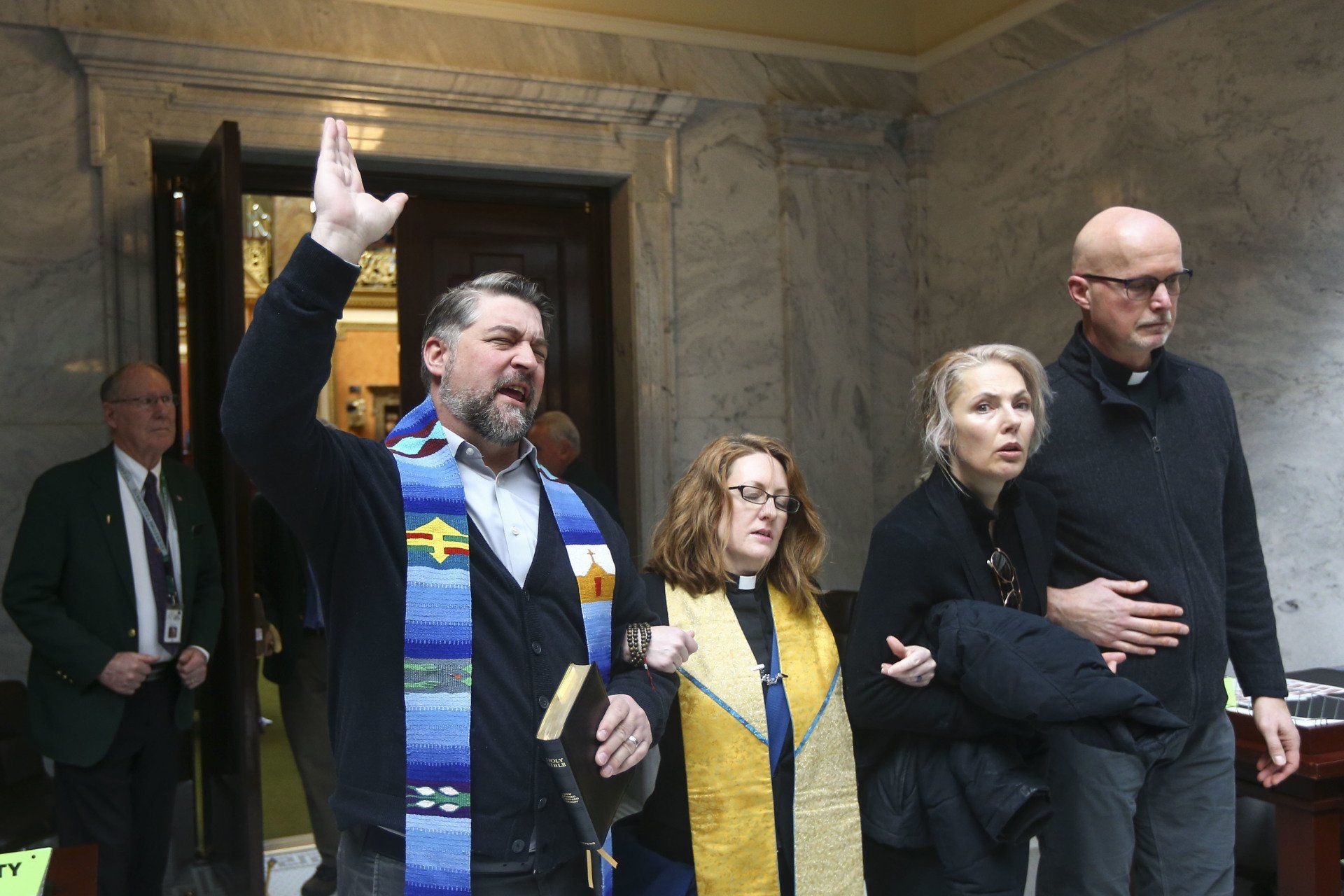 A man raising his hand, leading a group exiting a building; clergy attire.
