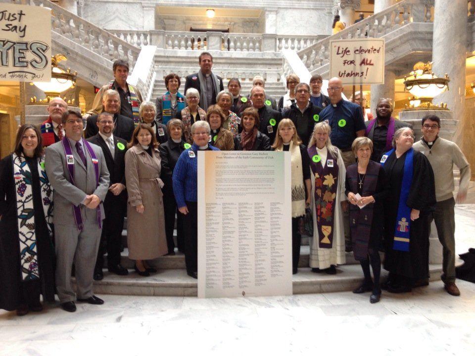 Group of people, some in clerical attire, stand with a sign in a building with a grand staircase.