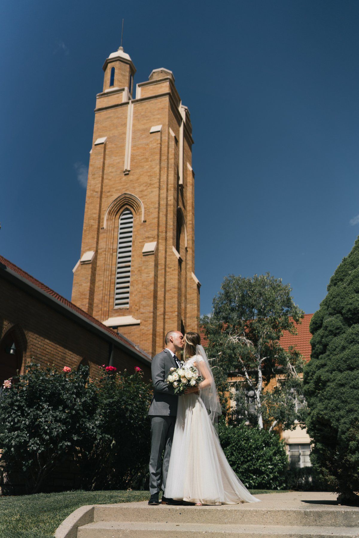 Bride and groom kissing in front of a tall brick church on a sunny day.