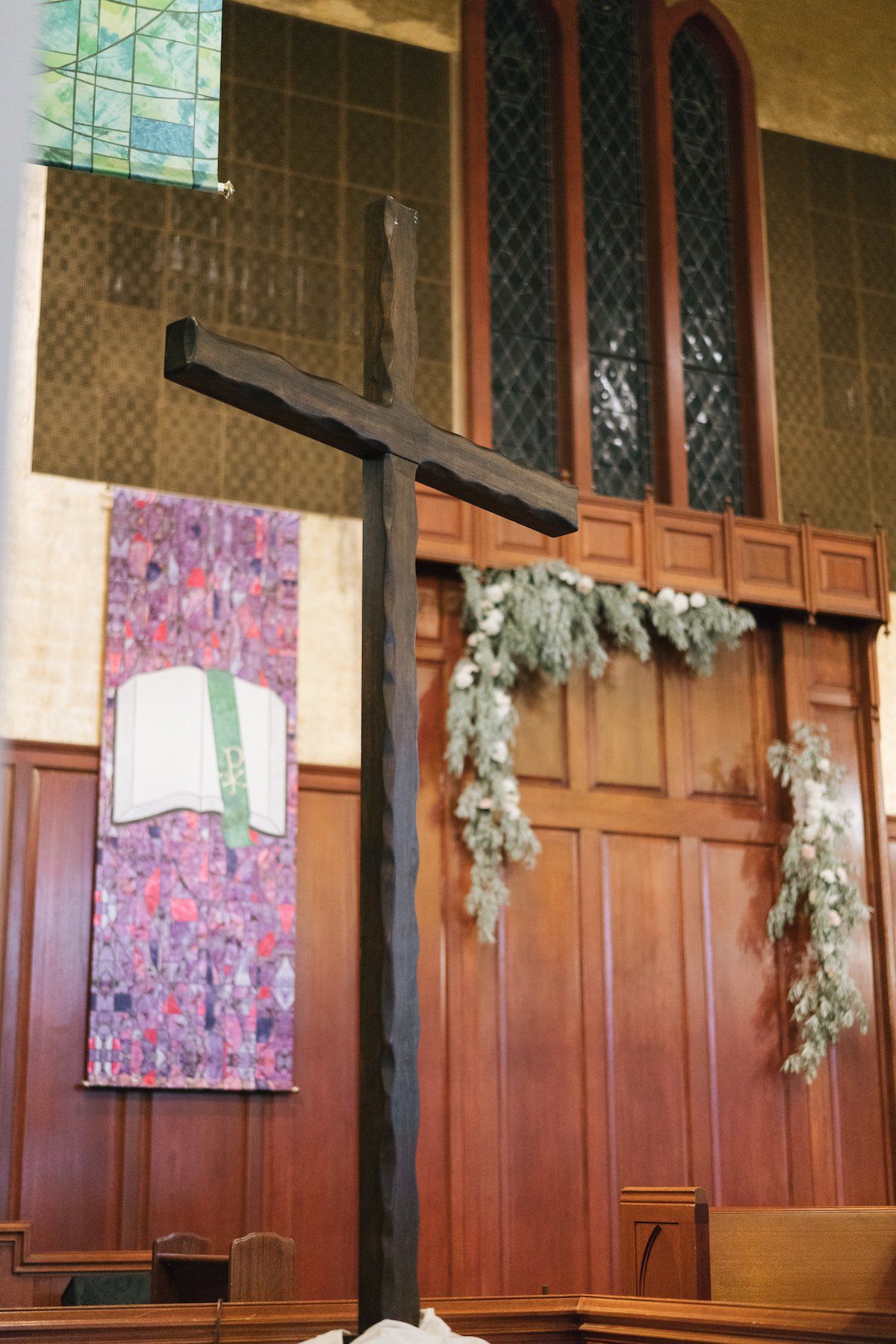 Wooden cross inside a church, in front of a wood-paneled wall, stained glass window, and hanging greenery.